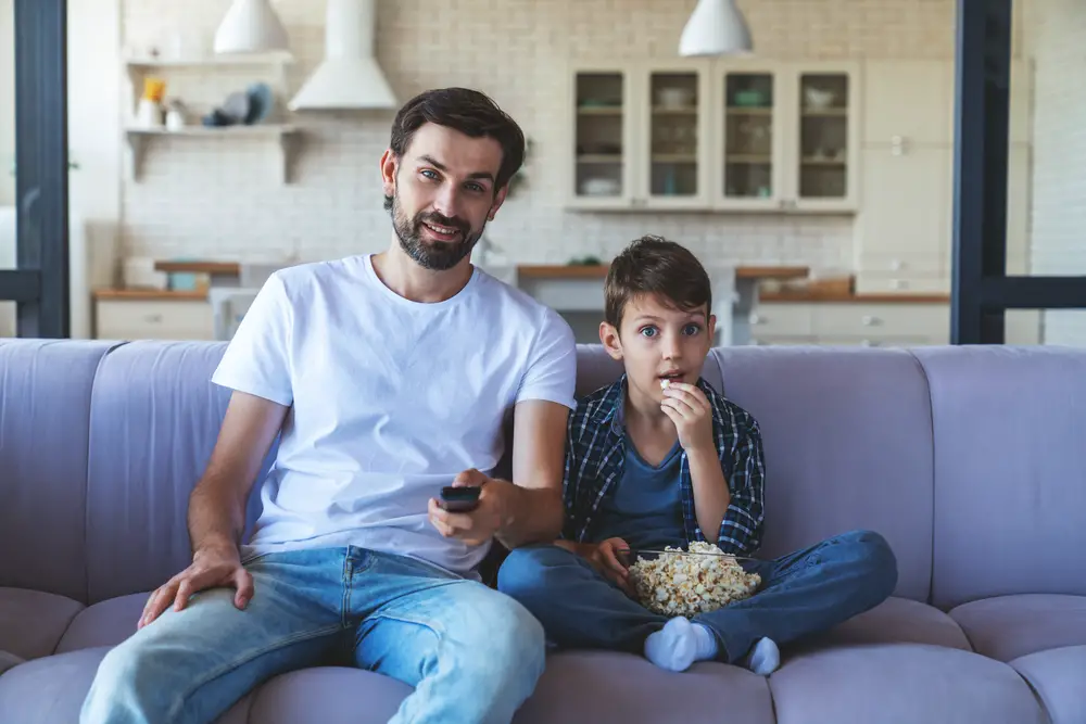 A happy little boy and his cheerful dad are sitting together on the couch in front of the TV, eating popcorn and cheerfully cheering for their favorite football team at home in the living room.